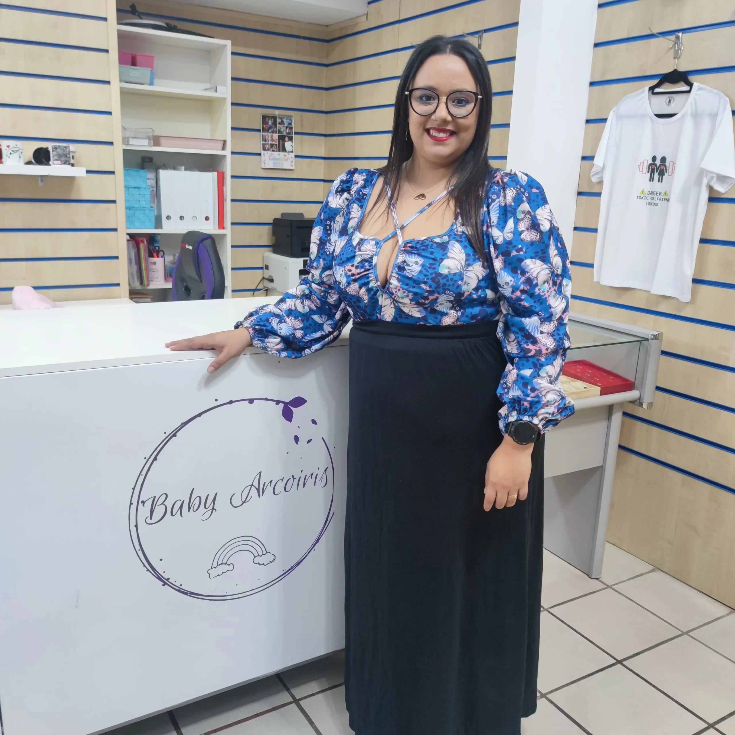 Mujer sonriente de pie junto al mostrador de "Baby Arcoíris", vistiendo una blusa azul con estampado de mariposas y una falda negra, en un ambiente de tienda que ofrece productos para bebés.